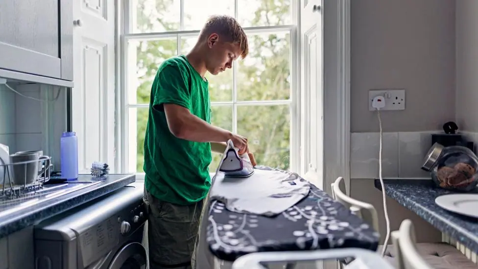 Stock photo shows a person ironing a T shirt in front of a window in their kitchen, next to a plug socket and with the washing machine behind them.