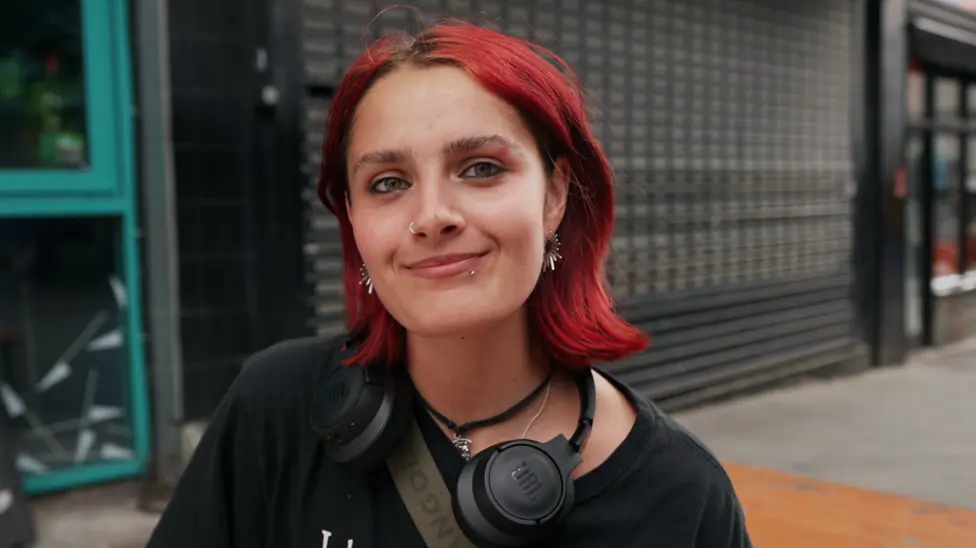 young woman with short red hair in the foreground, wearing a black T-shirt, black over-ear headphones around the neck, a cross-body strap, a nose ring and earrings. The person is facing the camera on…