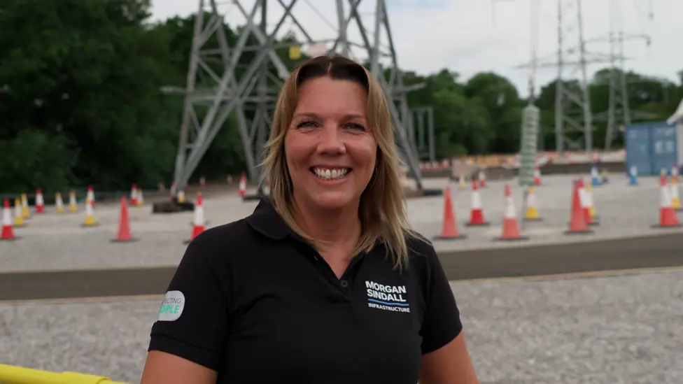 woman in the foreground wearing a black polo shirt with a “Morgan Sindall Infrastructure” logo, standing at an outdoor electrical infrastructure site and facing the camera. Behind the person are…