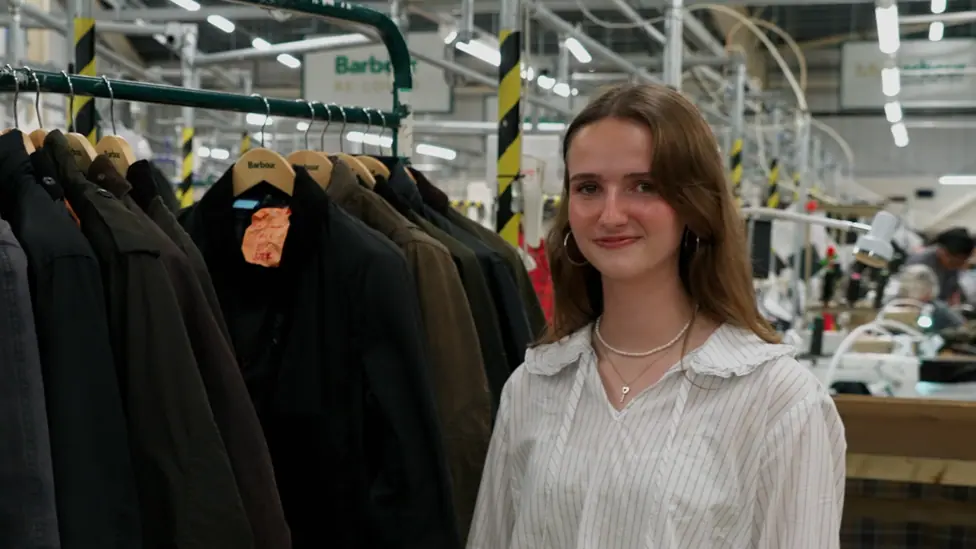 young woman standing in the foreground beside a clothing rail of dark waxed jackets on wooden Barbour hangers. The person is wearing a light striped shirt, hoop earrings and a necklace, and is facing…
