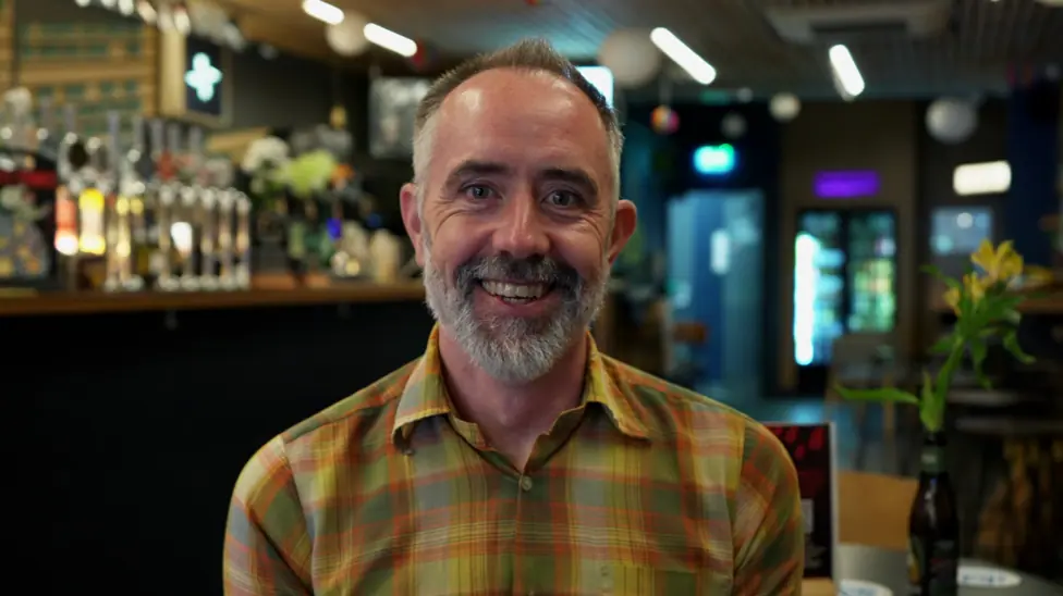 Man in the foreground wearing a yellow and green checked shirt, seated indoors and facing the camera. The setting is a bar or pub, with a row of beer taps and bottles visible behind the person on the…