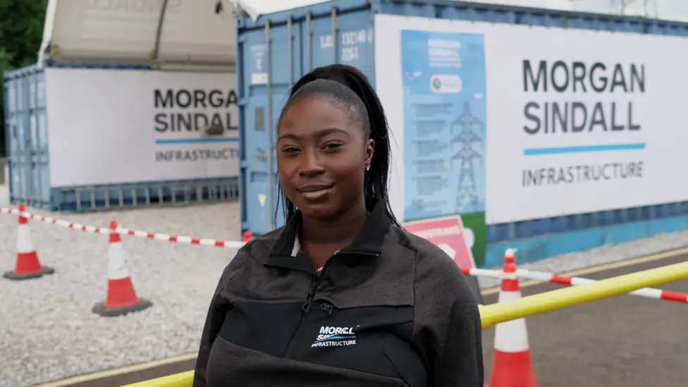 A young woman in a black Morgan Sindall Infrastructure zip-up jacket standing in the foreground at a construction or infrastructure site. Behind the person are two blue site cabins or container units…