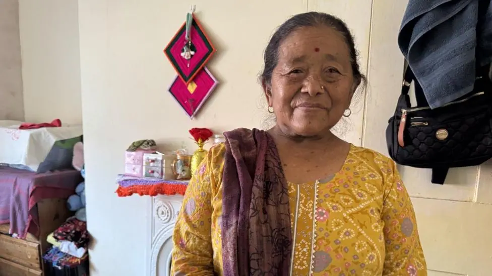 A Gurkha widow is wearing a bright yellow dress with a red shawl draped over her right shoulder. She also has a gold stud in her left nostril. Staring straight into the camera she's sitting in her…