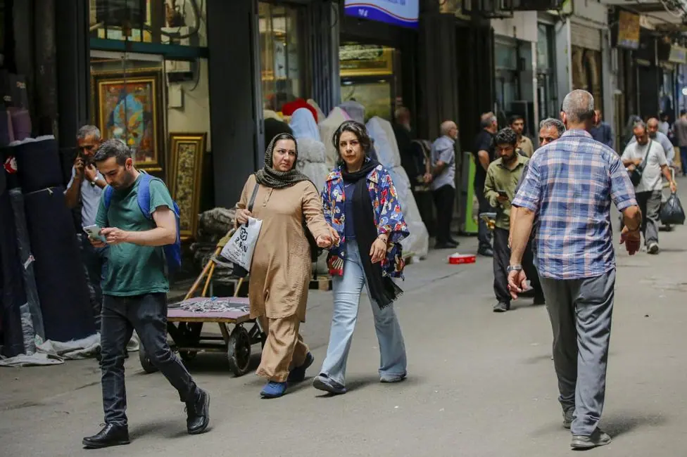 Several people walk on the streets of Tehran on a shaded street in July.