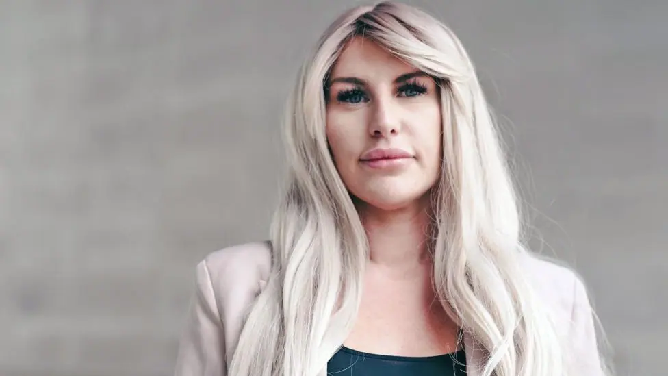 A head and shoulders portrait of Rebecca, standing against a light stone background. She has long blonde wavey hair and wears a black top with a light pink jacket. She looks pensively into the camera.