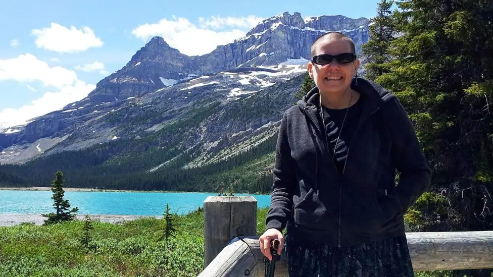 Cindy Baldassi is stood in front of a blue lake and towering mountain peaks with snow visible at highest points