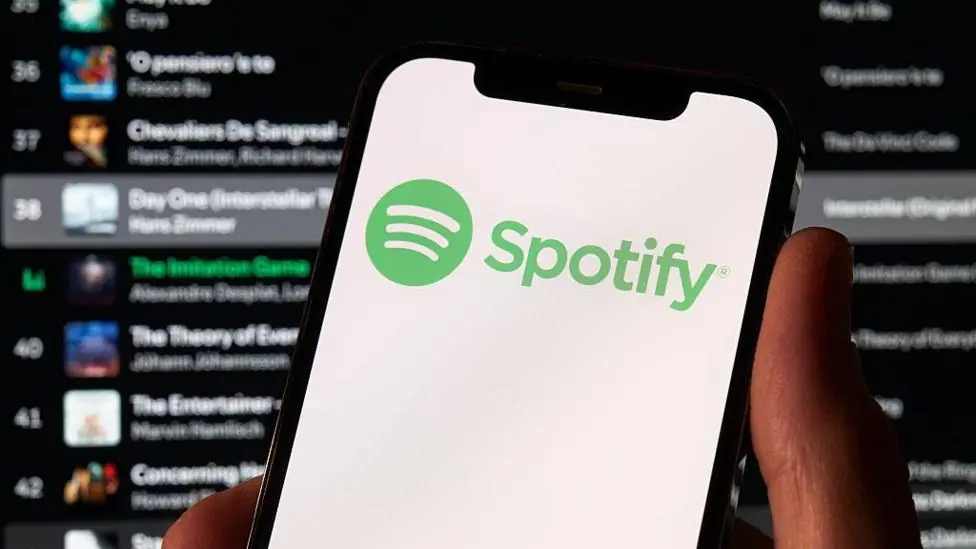 A photo of a hand holding a phone which has the green Spotify logo on it. Behind is a Spotify playlist in the background.