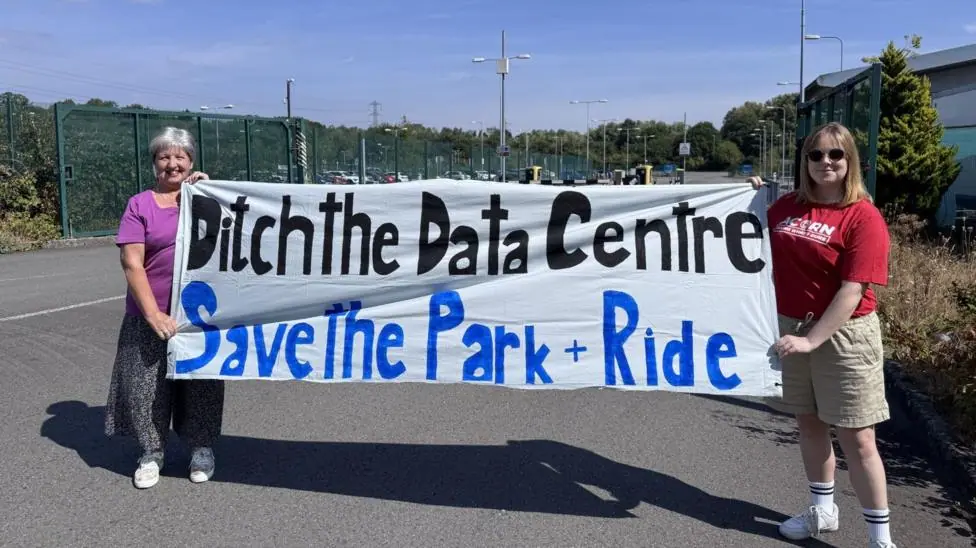 Two campaigners smiling holding a homemade banner which says "ditch the data centre, save the park & ride"