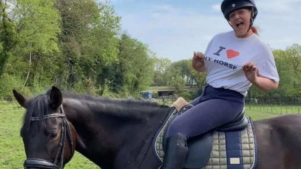 A woman, Tasha Lee Hopkins, sits on a horse pointing to her T-shirt, which reads: I love my horse. She is wearing dark blue jodhpurs, black riding boots and a black riding helmet. Her hair is tied…
