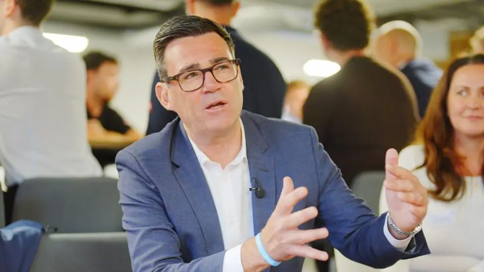 Andy Burnham gestures while speaking to people in a room. He is wearing glasses, a blue suit jacket, and a white shirt.