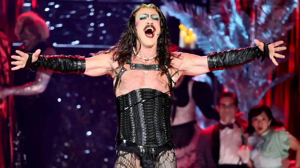 Luke Evans as Frank-N-Furter. He is wearing a black lace up corset, fish net tights and black leather gloves that go half-way up his arms. He is also wearing a wig and a full face of dramatic makeup.