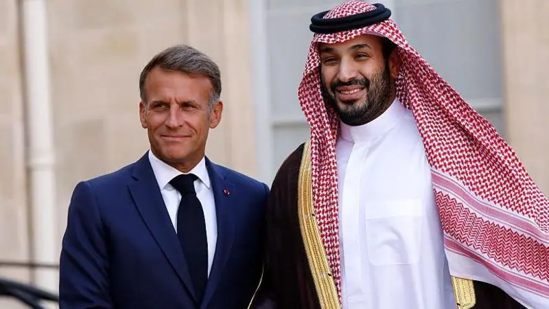 A smiling Macron, wearing a navy suit and black tie, poses next to bin Salman, who is wearing traditional Saudi attire