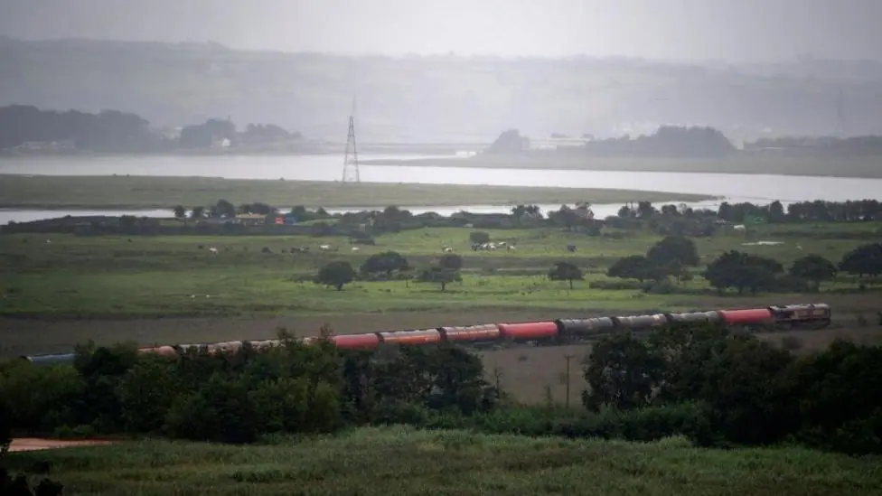 A train travelling through a rural area with a river and marshes seen in the background.