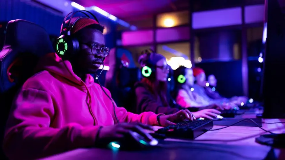 A man is playing a video game on a computer, there are other gamers, wearing the same green illuminated headphones, out of focus along the desk to his left.