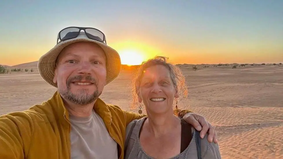 A selfie taken by Nick Smith showing him wearing a yellow jacket and a beige undershirt, and a beige hat with sunglasses sitting on top of his head, his arm around his wife, who is smiling and has an…