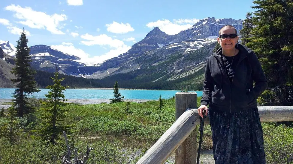 Cindy Baldassi is in the foreground wearing sunglasses and a dark hoody and navy blue dress. She is looking at the camera and behind her is a beautiful blue lake and towering mountain peaks with snow…