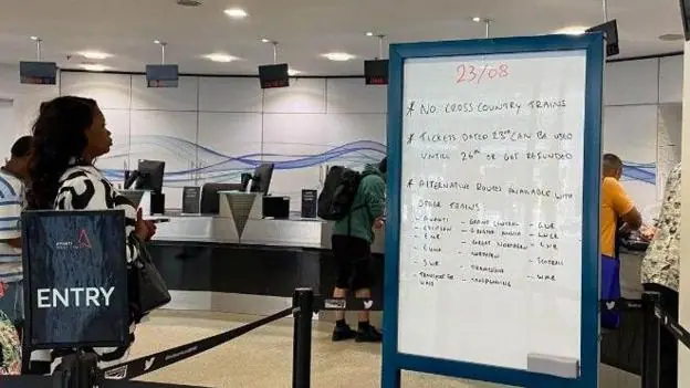 A hand-written whiteboard details the cancellations to trains, in the customer ticket office. On the right there is a sign that says Entry, behind which passengers are waiting to go to the counter.
