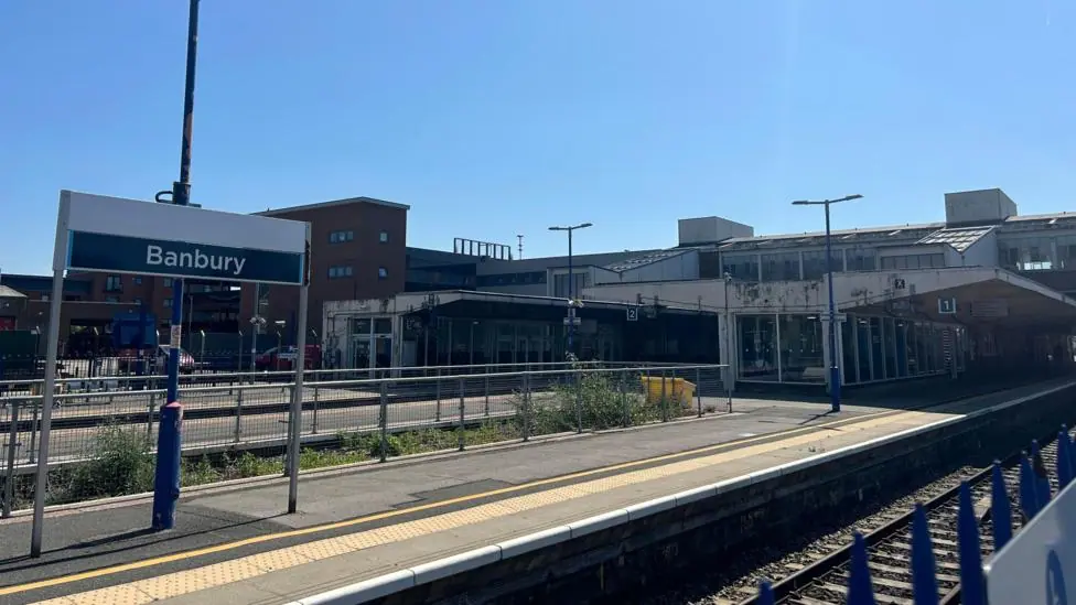 A sign saying Banbury sits above a train platform and railway line in the foreground, further platforms are visible in the distance. In the distance, an ageing building is also visible - this is the…