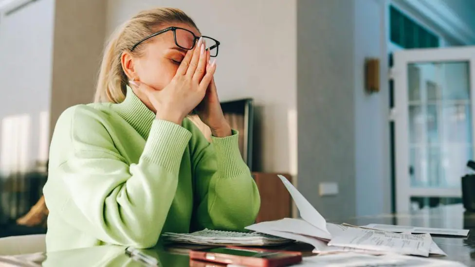 Woman sits at a table at home with her head in her hands, with paperwork in front of her.