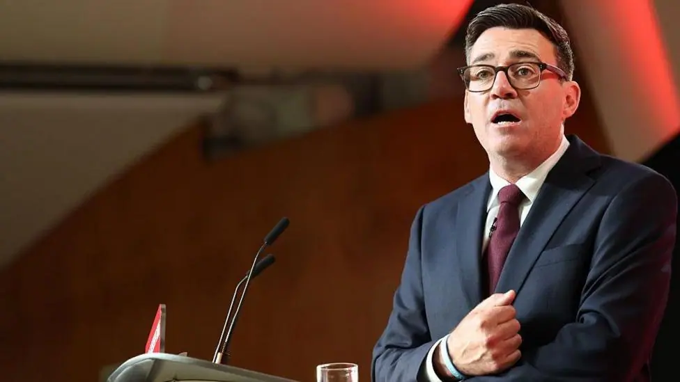 Andy Burnham stood at a lectern delivering his first speech as the leader of the Labour Party. His right hand is clenched in a fist and pulled towards his chest as he speaks. He is wearing glassed, a…
