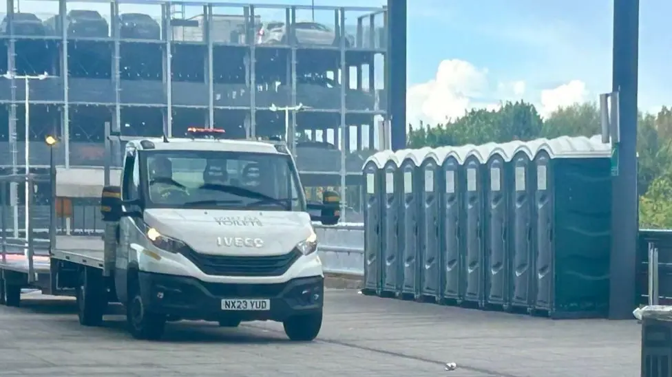 A vehicle is parked next to a line of portable toilets, with a multistorey carpark in the background