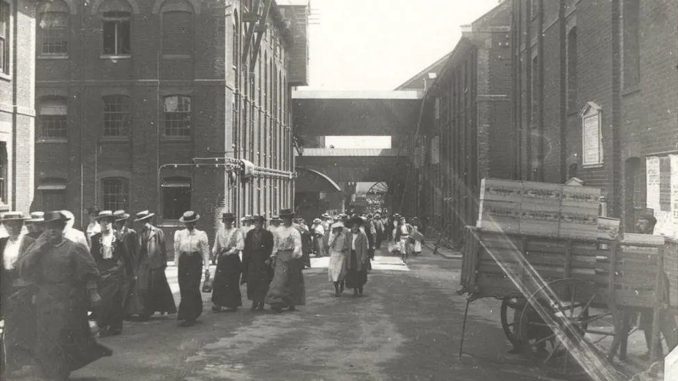 A black and white photograph believed to be from 1910 of Colman's workers leaving a factory. A large crowd of people wearing hats are walking from the site towards the left of the frame.