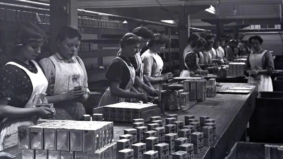 More than 14 people standing in a row at a table as part of a production line inside Colman's. The picture is black and white and is believed to have been taken in 1908.