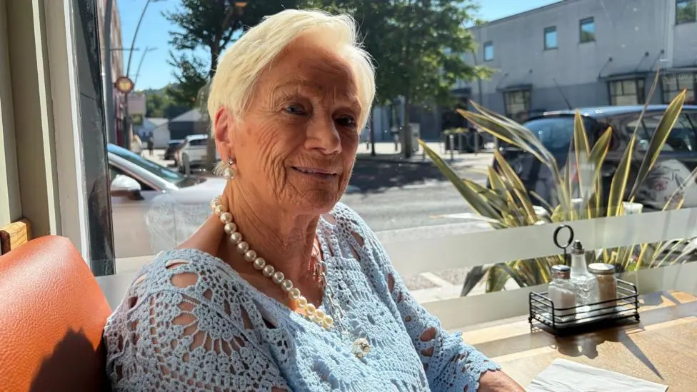 Isobel Russell. She has short blonde hair, wearing pearl earrings and necklace and light blue lace top. She is sitting at a cafe table by a window.