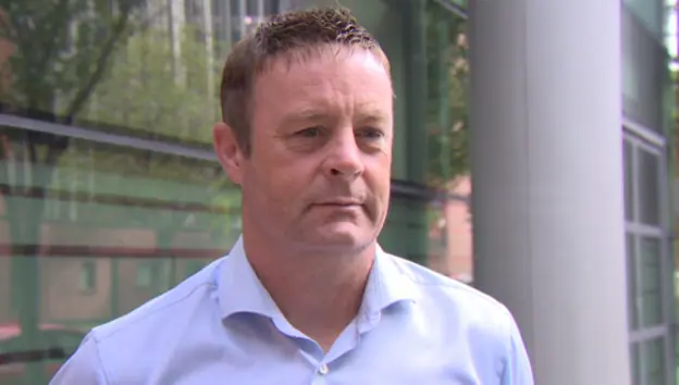 Raymond Gormley has short brown hair and is wearing a white shirt. He is standing near a large building with a number of windows.