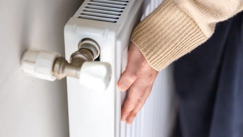 Someone is holding their hand against a radiator. Their face cannot be seen. The radiator is white and the person is wearing a yellow jumper and navy bottoms.