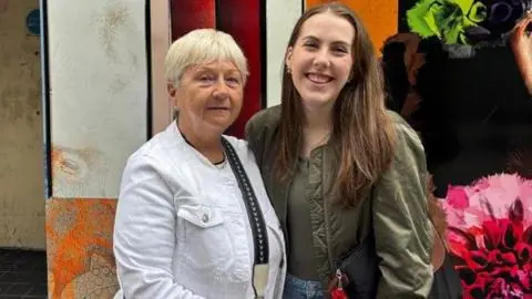 On the left is Agnes, wearing a white jacket, black jeans with a black and cream cross body bag. She has short blonde hair. Beside her on the right is her granddaughter Abbie, wearing blue jeans, a…