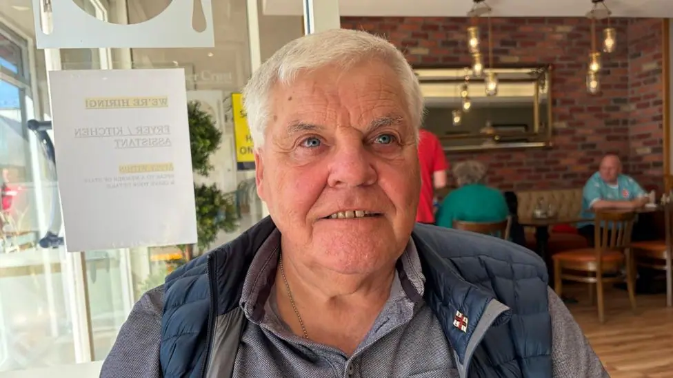Gawn Graham. He has short white hair, wearing a navy puffer gilet and grey polo shirt. People are sitting at cafe tables behind him.