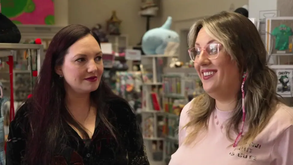 Two women in a shop. One has dark hair and red lipstick and the other has blonde hair and pink glasses.