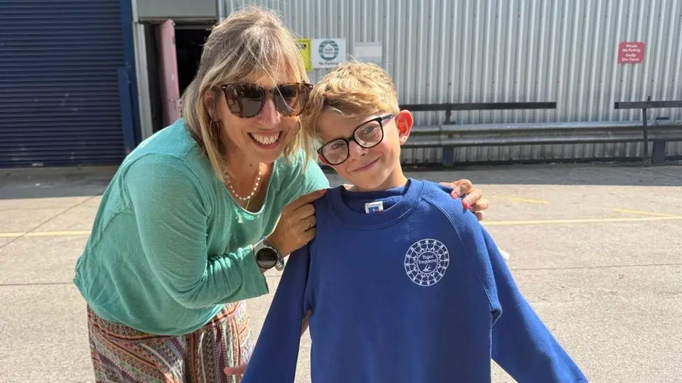 Elise wears a green top, and multi coloured trousers and has blonde hair and wears sunglasses. She is pictures next to her 10 year old son, Charlie, holding a blue jumper next to which she got from…