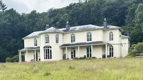 Dol-llys Hall taken from the far side of a long-grass lawn. The hall is painted light yellowy cream and has a slate tiled roof. To the left and right of a round tower-like protuberance are two…