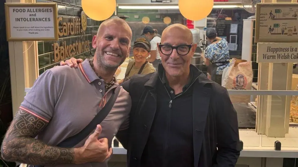Owner of the Welsh cake store stood next to actor Stanley Tucci. He has a full sleeve tattoo and a bag strap across his body. Tucci is wearing a black jacket over a black quarter zip jumper. There is…