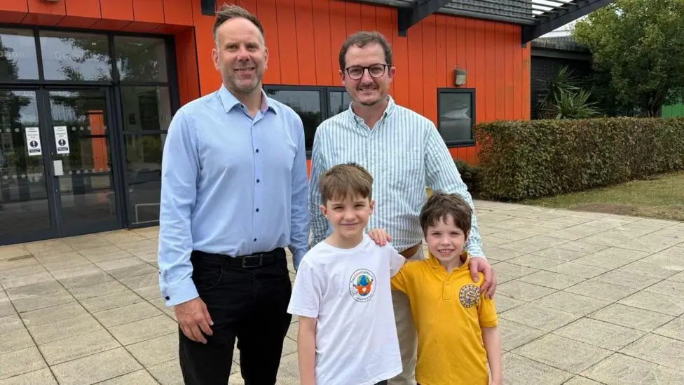 Deputy head teacher, Gareth Evans wears a blue shirt and black trousers and is pictured next to Jon who is wearing a striped shirt and cream trousers and his two children. 10 year old, Tom wears a…