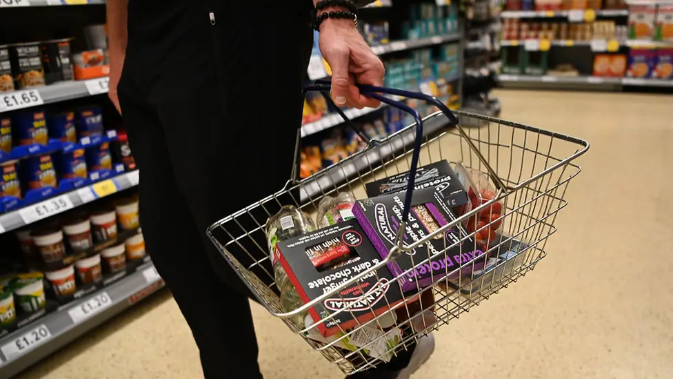 Person holding a wire shopping basket in a supermarket aisle, containing packaged groceries including chocolate desserts, salad, and other food items, with store shelves stocked with products in the…