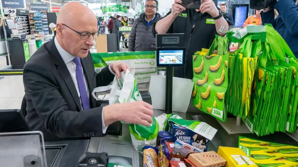 John Swinney is sitting at an Asda checkout where he is wearing a dark suit, white shirt and purple tie. He is bagging a pile of shopping as photographers and camera people capture the image.