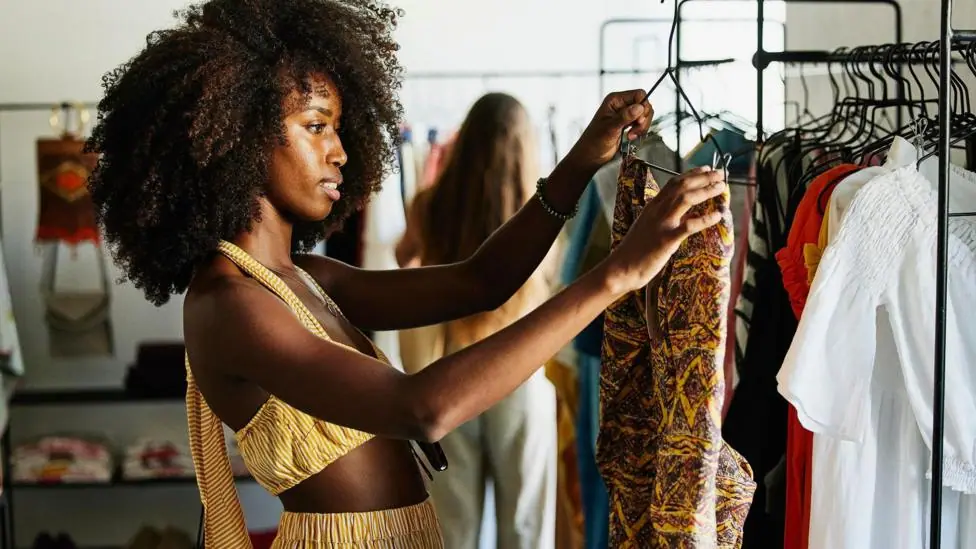 A shopper inspects a a vibrant, patterned outfit she has picked out from a clothes rack. She is dressed in a striped yellow outfit.