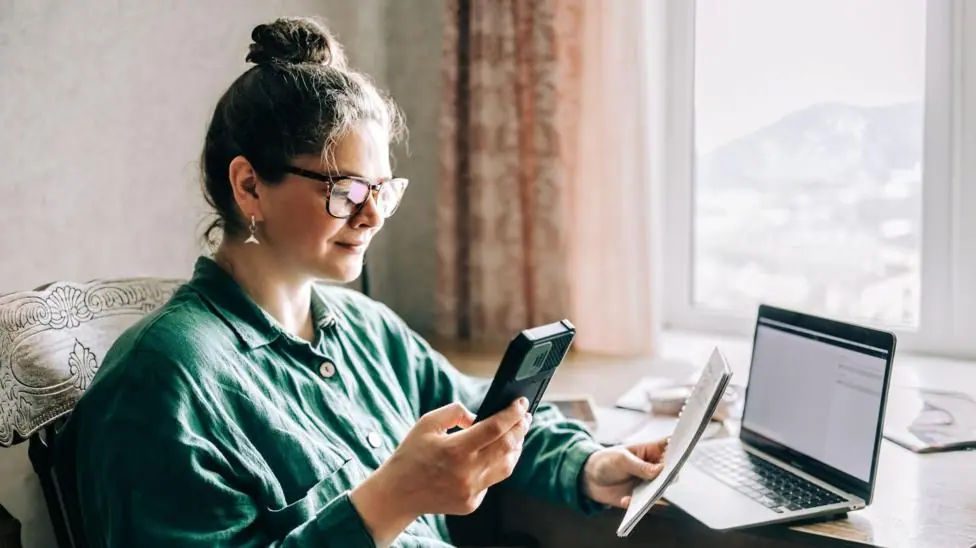 Woman sits at a window seat holding a phone and a pad with a computer on a table in front of her.