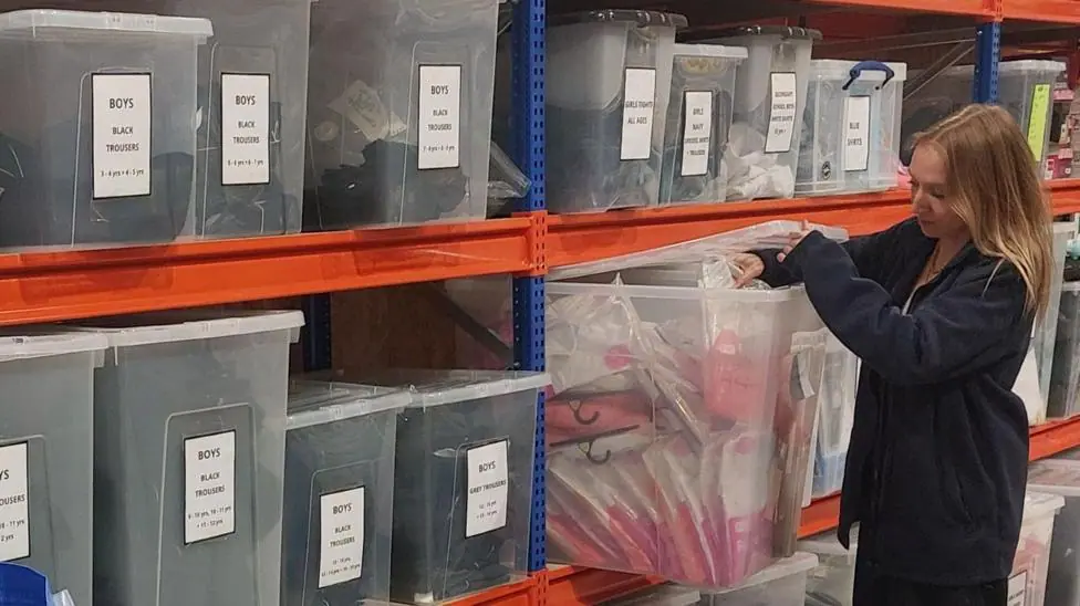 A volunteer stands next to boxes of boys' black and grey school trousers.