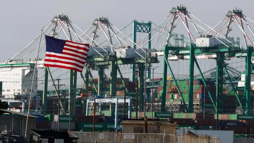 An American flag waves against a backdrop of cranes and storage containers in Los Angeles.