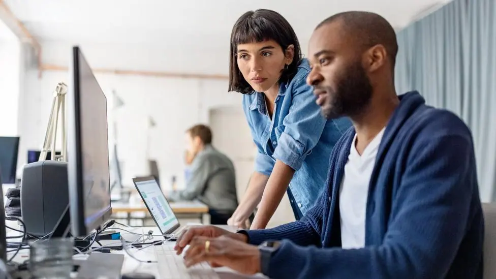 Stock photo shows a person leaning over the computer of another person in an office environment