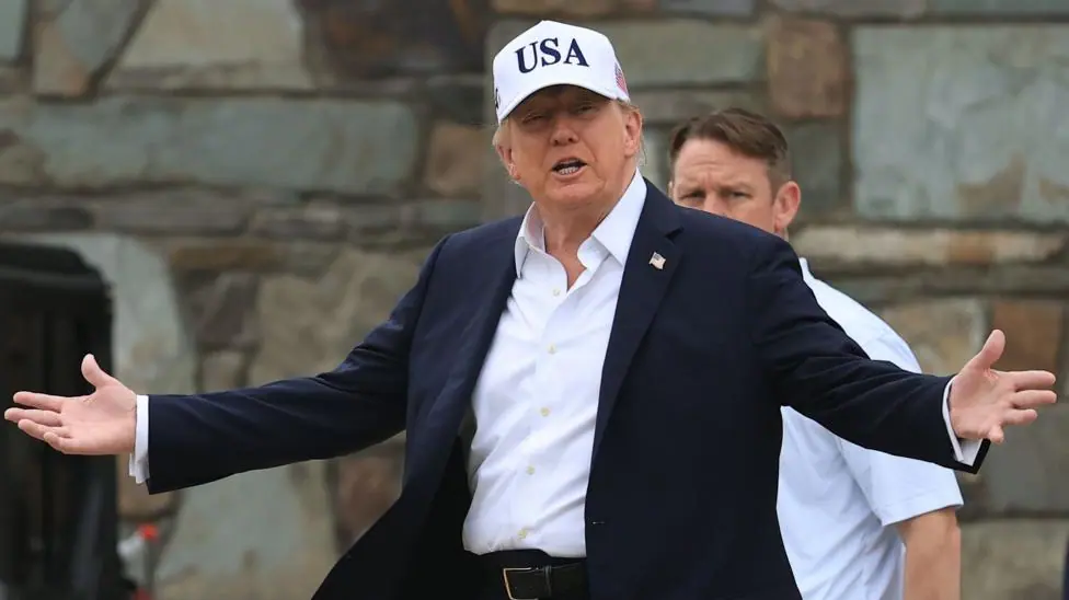 U.S. President Donald Trump attends the Amgen Irish Open at Trump International Golf Links in Doonbeg, Ireland. He is wearing a white USA baseball cap, navy blazer with a white shirt. He has his arms…