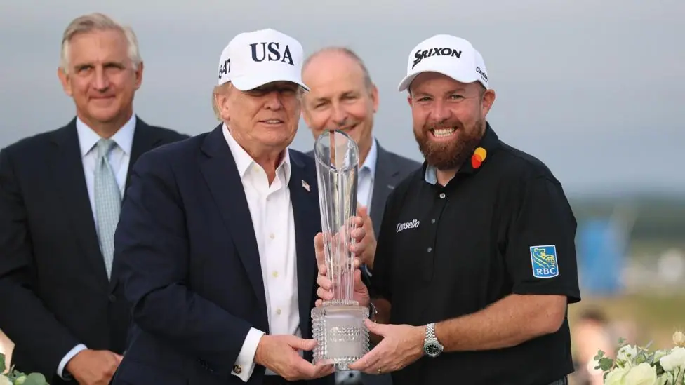 Two men stood, holding a trophy. They are both smiling. On the left is Donald Trump, wearing a navy suit and a white hat with 'USA' written on it. On the right is Shane Lowry, wearing a black polo…