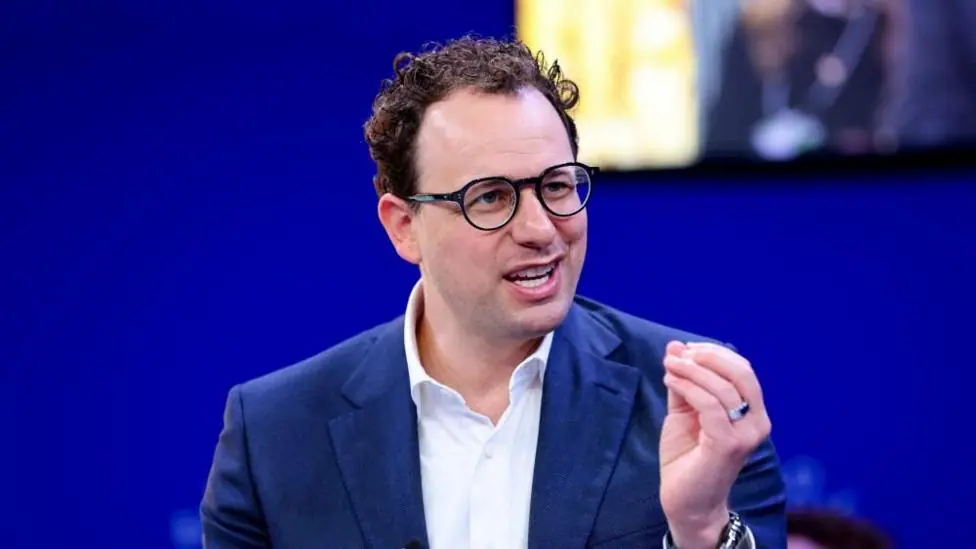 Dario Amodei hld the closed fingers of his left hand together as he mades a point. He is wearing a white shirt and blue jacket, black-rimmed glasses and has slightly curly hair. He is sat against a…