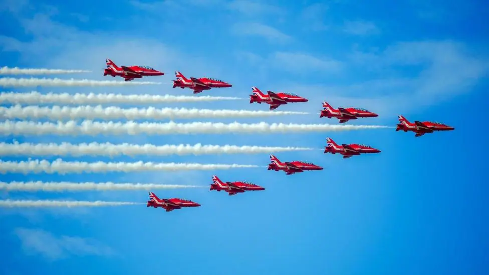 Nine red planes fly in formation through the sky. A trail of white smoke follows each of them. The aircraft are flying in an arrow shape against the backdrop of a brilliant blue sky.
