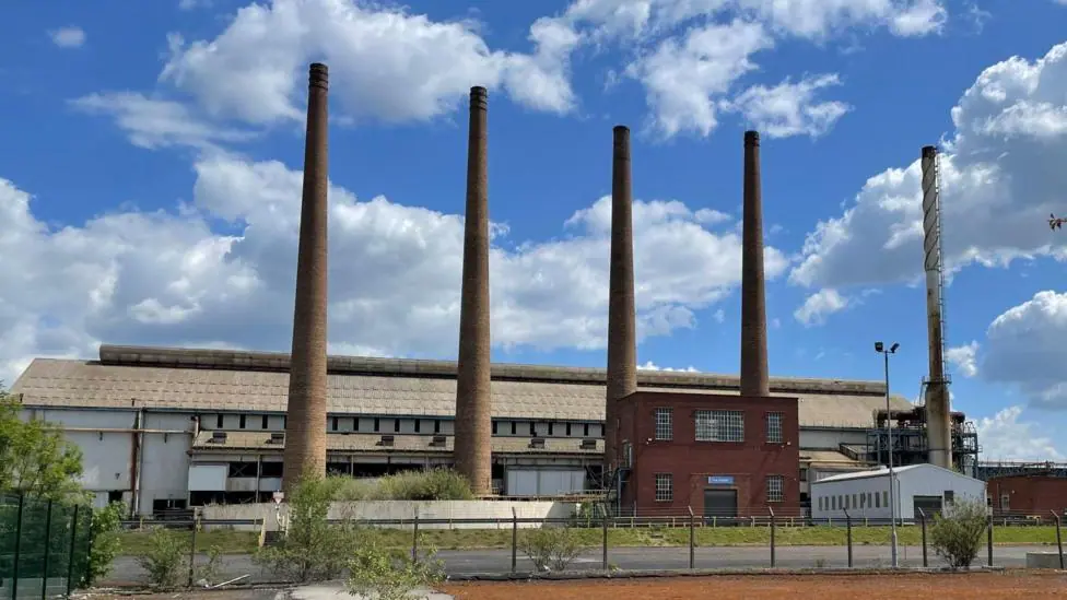 A factory with five tall narrow chimneys rising from the ground. The sky is blue with some white clouds floating above.