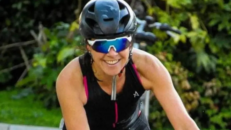 Rebecca Comins is wearing a black and pink tri-suit, she is riding her bike. She is wearing sunglasses and a helmet and smiling